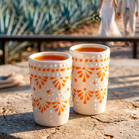 Two white ceramic tequila shot glasseswith orange floral patterns sit on a stone table in an outdoor agrotecho setting, with a bride and groom blurred in the background walking along a path through an agave field.