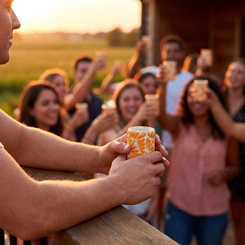 A man in a blue t-shirt leans on a wooden railing at sunset, holding a patterned orange and white talavera shot glasses, with a group of friends and family celebrating on a porch in the background.